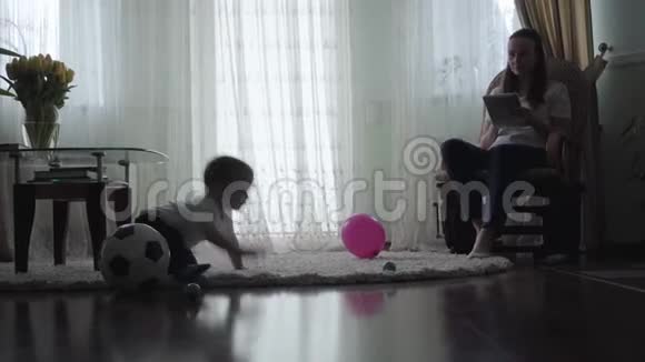 可爱的婴儿在地板上爬行在蓬松的地毯上玩球和气球而他的年轻母亲坐在里面视频的预览图
