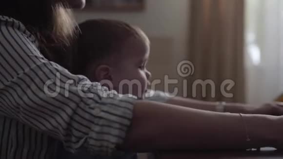 小儿子坐在母亲的腿上在桌子上靠近女人的手打开笔记本电脑忙碌的年轻母亲在家工作视频的预览图