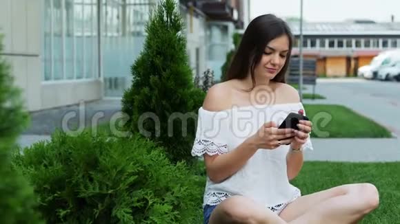 年轻漂亮的女人坐在酒店附近的草坪上用电话打印一条信息视频的预览图
