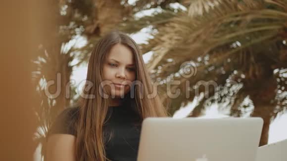 漂亮的女学生或在笔记本电脑上工作的自由职业者坐在阳台上看棕榈和大海速度很快视频的预览图