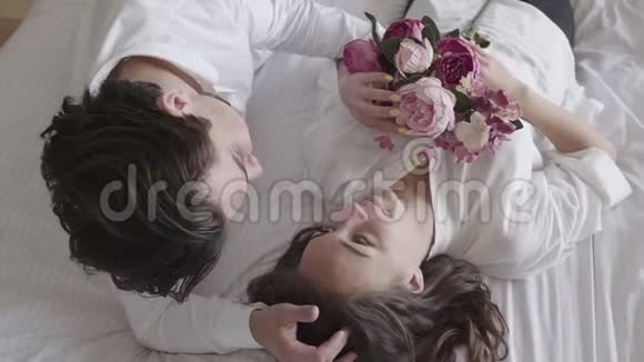头顶拍摄幸福的年轻夫妇躺在床上说话女人抱着花束微笑的特写投标关系视频的预览图