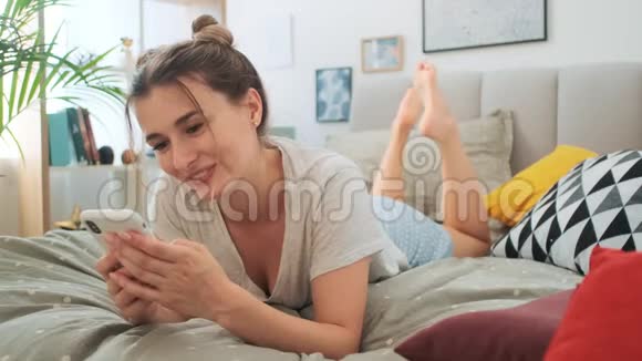年轻的白种人女孩在床上休息时一边在智能手机上发短信一边打字一边兴高采烈地微笑着视频的预览图