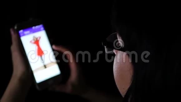 一个戴着眼镜的深色头发的女孩在晚上用手机特写通过一家网上商店看网上购物视频的预览图