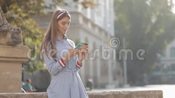 美丽的年轻女子穿着蓝色和白色条纹连衣裙衬衫站在老街上使用智能手机和智能手机视频的预览图