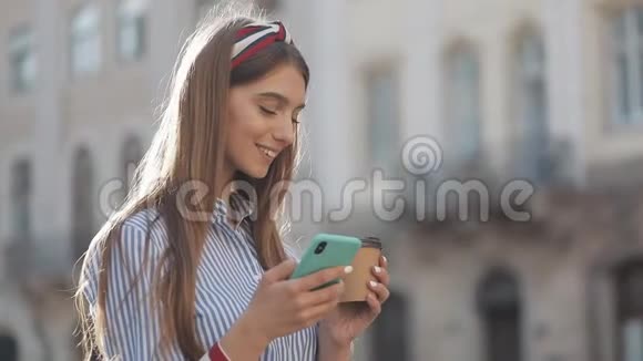 美丽的年轻女子穿着蓝色和白色条纹连衣裙衬衫站在老街上使用智能手机和智能手机视频的预览图