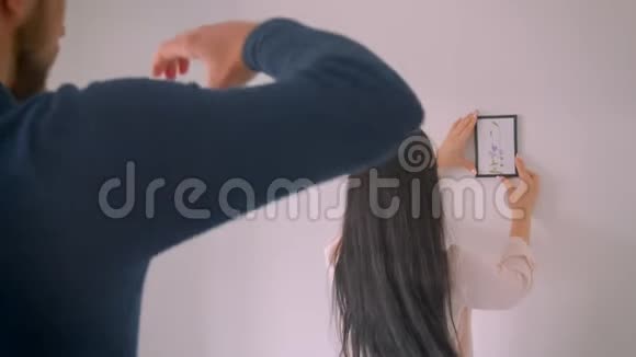 白种人的女孩正在选择白色墙壁上的相框图片的位置而她的男朋友用手指做相框形状视频的预览图