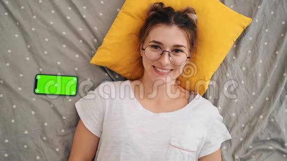 躺在床上的戴着眼镜的年轻迷人女孩的俯视图中她兴高采烈地对着镜头笑着用一个视频的预览图