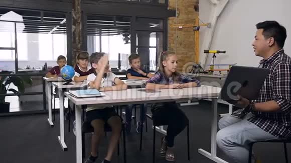 可爱的白种人男孩和同学坐在桌子后面举手向韩国老师提问视频的预览图