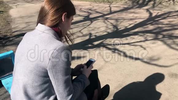 一个穿着灰色外套的女孩坐在长凳上用手机看风景视频的预览图