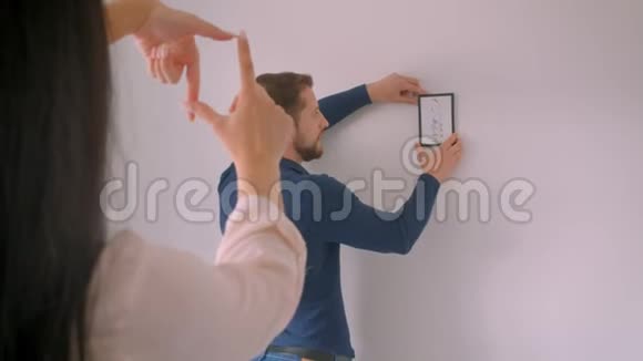 白种人的男人选择了在白墙上画框的位置而他的女朋友用手指做框的形状视频的预览图