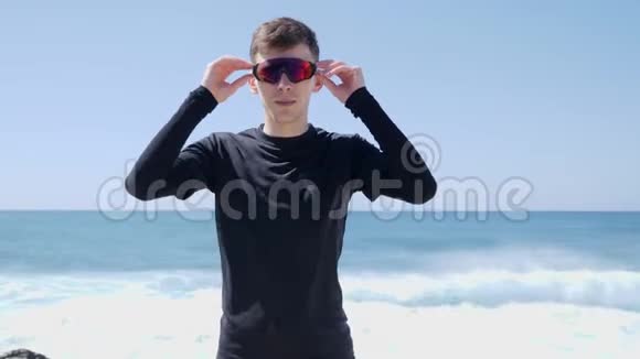 年轻自信的运动员脱下太阳镜站在海滩上身后有波浪晴天视频的预览图