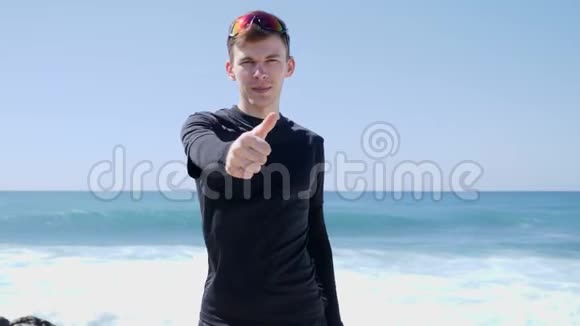 年轻的身材英俊的黑衣人竖起大拇指对着站在海滩上的摄像机强烈的海浪在撞击Bea视频的预览图