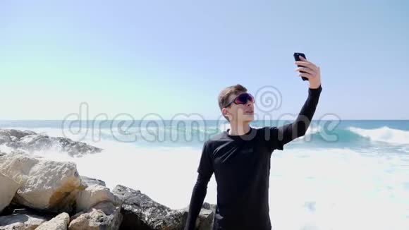 年轻的身材健壮的白种人穿着墨镜拿着电话站在岩石海滩上带着自拍视频的预览图