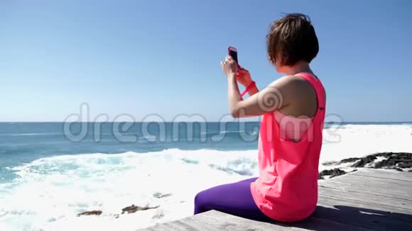 年轻美丽的高加索女人坐在岩石海滩上拿着电话拍照慢动作视频的预览图