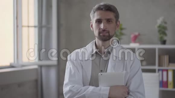 英俊的技巧微笑的男人穿着白色长袍看着相机拿着平板电脑现代成人医生视频的预览图