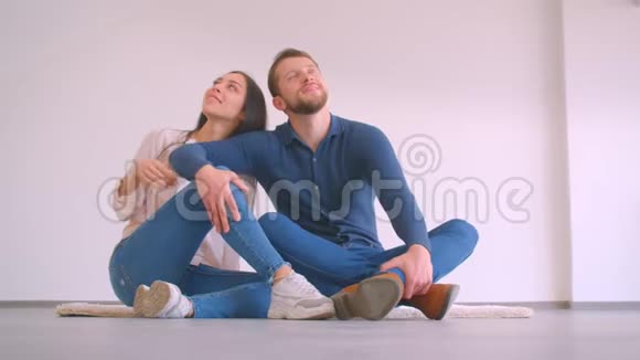 年轻的白种人夫妇坐在新的空的地板上平抱亲吻在白色的墙壁背景下快乐视频的预览图