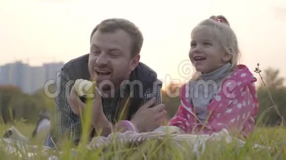 白种人的父亲和他的金发小女儿坐在草地上吃苹果的肖像小女孩视频的预览图