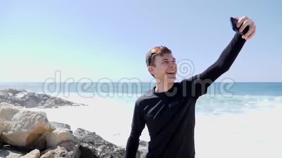 年轻快乐的微笑着的男人在岩石海滩上拍自己的照片笑着竖起大拇指溅起浪花视频的预览图