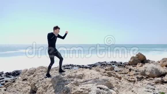 年轻苗条的男子拳击空气在岩石海滩强浪正在撞击海岸风和日丽的日子视频的预览图