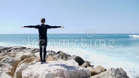 年轻的白种人站在岩石海滩上手臂张开强烈的波浪撞击悬崖和水溅阳光明媚视频的预览图
