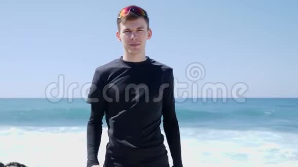 年轻的身材英俊的黑衣人竖起大拇指对着站在海滩上的摄像机强烈的海浪在撞击Bea视频的预览图