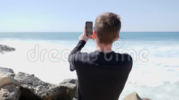 年轻的帅哥在一个阳光明媚的风日里拍着海洋和海浪拍打着岩石海滩的照片溅水花关闭视频的预览图