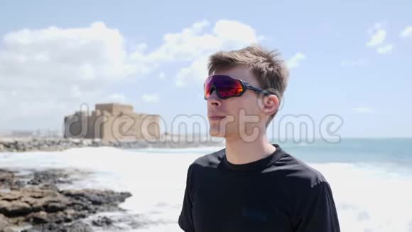 戴太阳镜的年轻运动员在训练前戴上耳机站在海滩上波浪正在撞击海岸线视频的预览图