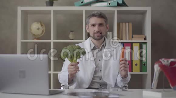 一幅英俊的积极男子的肖像穿着白色长袍在镜头中提出胡萝卜和西兰花熟练的医生视频的预览图
