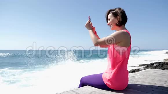 年轻美丽的白种人女人坐在岩石海滩上拿着电话拍着海浪溅到岩石上的照片视频的预览图