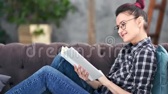 微笑的时髦女孩戴眼镜看书坐在家里的沙发中弹视频的预览图