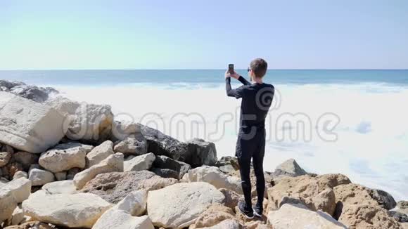 年轻的帅哥在一个阳光明媚的多风天气里拍着海洋和海浪拍打着岩石海滩溅起水花慢慢慢慢视频的预览图