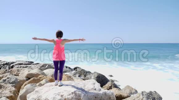 年轻的白种人黑发女孩站在岩石海滩上双臂张开强烈的海浪拍打着悬崖溅起水花视频的预览图
