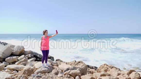 年轻漂亮的适合女孩拿着电话和自拍照片而站在岩石海岸波浪在岩石上飞溅慢慢视频的预览图