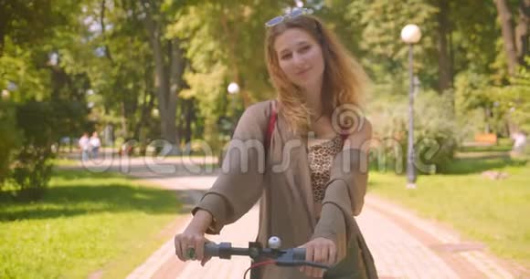 在公园里年轻漂亮的白种人女性站着踢滑板车看着镜头开心地笑着视频的预览图