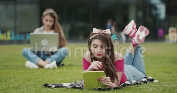 两名女性坐在一个可爱的公园的草地上使用笔记本电脑和平板电脑两个漂亮女孩视频的预览图
