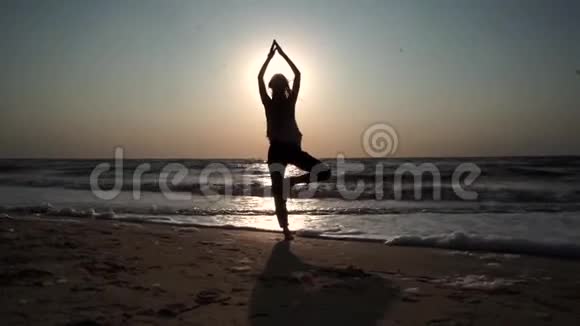 女孩站在瑜伽姿势上日落时太阳对面海滩上大海上视频的预览图