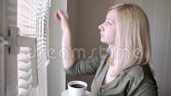 早晨站在窗前的一个年轻的忧郁的美丽的金发女人喝着咖啡举起百叶窗视频的预览图