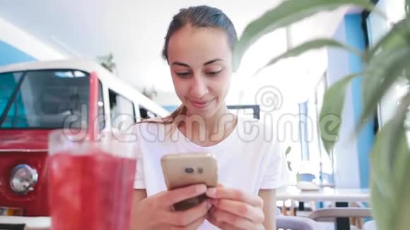 穿着白色t恤的漂亮年轻晒黑的女人坐在咖啡馆里用电话看读或搜索什么东西视频的预览图