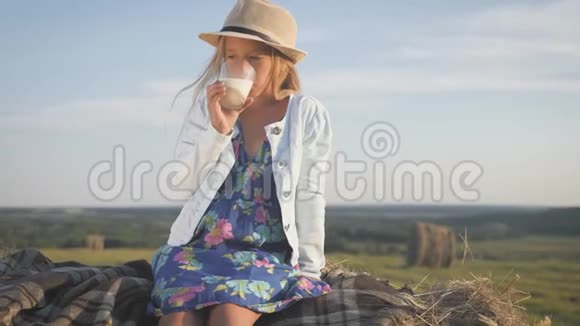 穿着蓝色裙子的漂亮女孩在田里的干草堆上喝牛奶孩子在夏天野餐视频的预览图