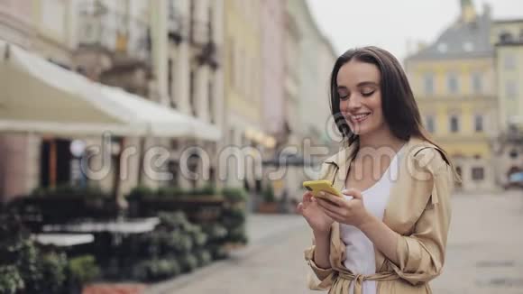 迷人的时尚少女使用她的智能手机打字和微笑站在旧城背景视频的预览图
