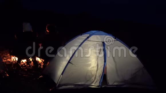 一对年轻夫妇坐在火炉旁在海边用帐篷露营手持设备的背面视图视频的预览图