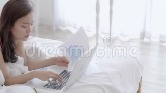 漂亮的亚洲自由职业女性在卧室的床上用笔记本电脑工作和打字视频的预览图