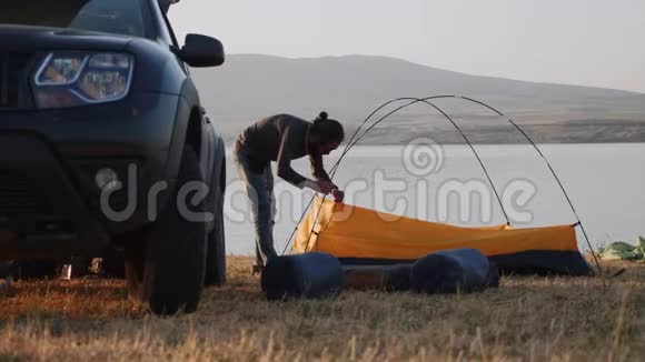 一个留胡子的年轻人在靠近他的车的湖岸的露营地搭起一个帐篷视频的预览图