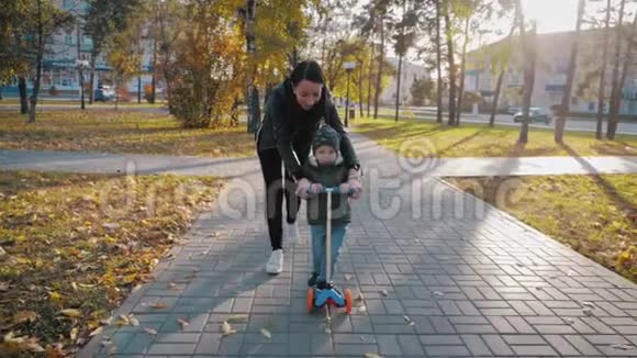 年轻的母亲教她蹒跚学步的儿子如何在秋天的公园里骑滑板车积极的家庭休闲培训体育视频的预览图
