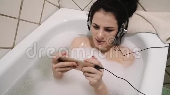 年轻女孩用防水智能手机耳机听音乐在泡泡浴中唱歌视频的预览图