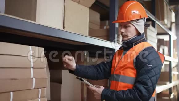 戴着头盔和工作服的胡子和胡子的工人站在架子旁边数着ipad上的工具视频的预览图