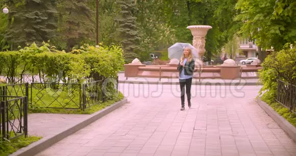 美丽的白种人金发学生带着雨伞沿着绿色公园散步观察大自然的美丽视频的预览图