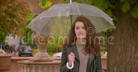 在绿色城市公园里白种人深色头发的女性带着雨伞微笑美丽而快乐地进入镜头视频的预览图