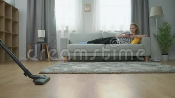 疲惫的丈夫在地板上吸尘妻子在沙发上玩智能手机视频的预览图