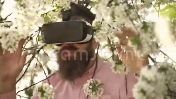大胡子男人戴着VR眼镜在盛开的花园里享受阳光明媚的一天穿着精致白花的英俊男子视频的预览图